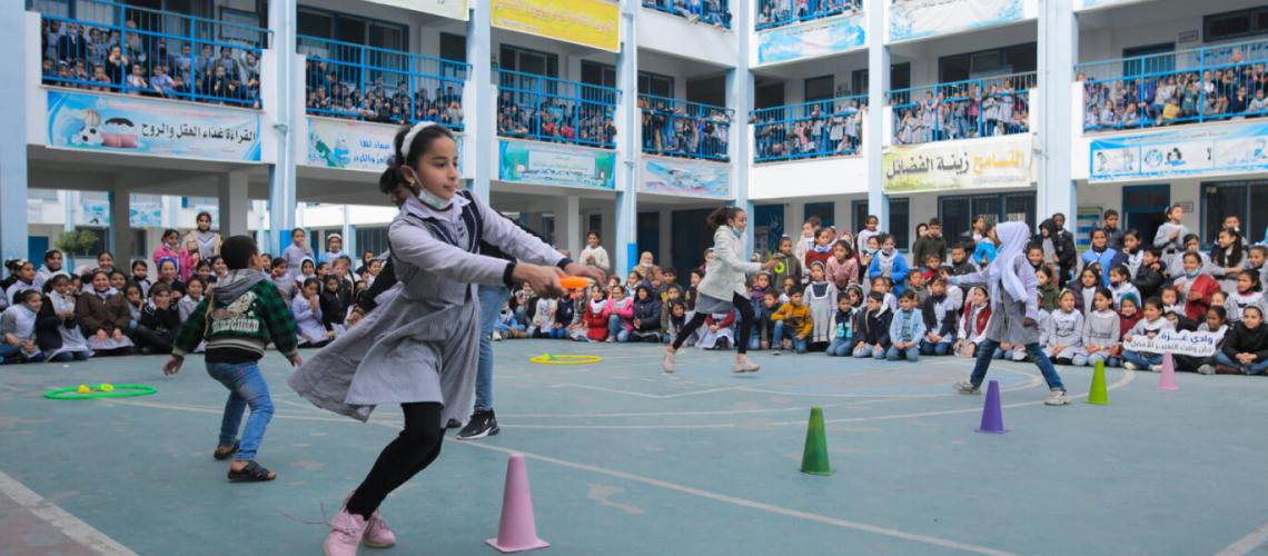 Kids play festively on a soccer/tennis court inside an open-air courtyard of a school painted white with blue trim.