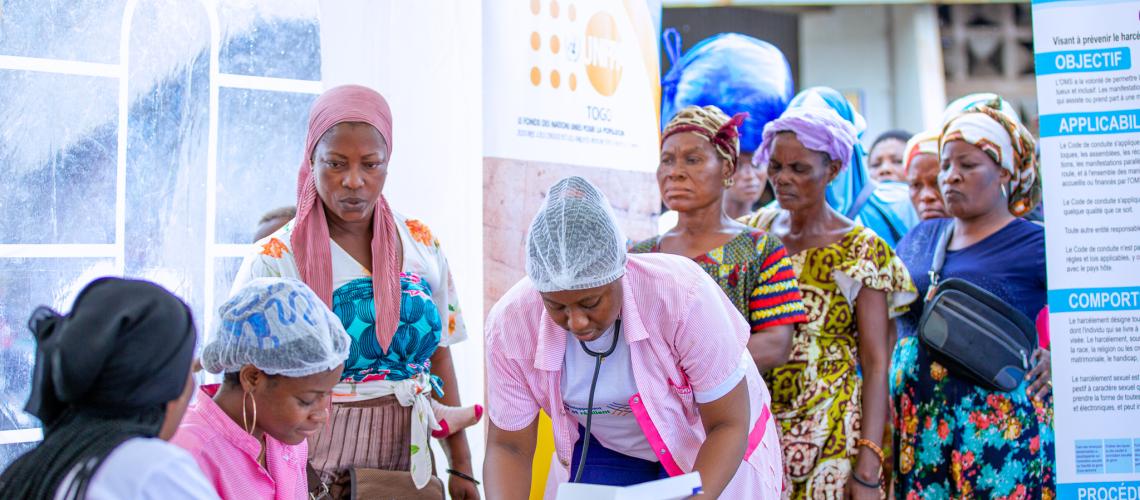 A group of women, some of them in pink doctor or nursing uniforms are bent over a table, appearing to be writing something. A line of women in colourful clothes and headscarves are standing behind the table waiting to be attended to.